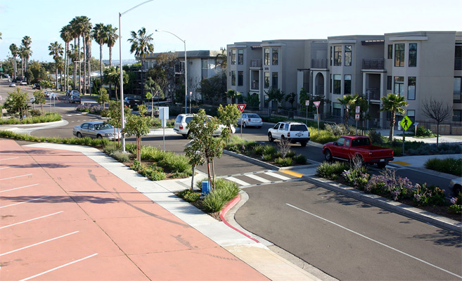 an arial view of the same street shows that the intersection has been replaced with a roundabout and the crosswalk moved about a half block away.  At the crosswalk, the middle lane has been replaced with a planter, the right side of the street has been narrowed to only one lane, and sidewalk on the left side of the street extends into the street and blocks the parking lane, making the crossing only one lane to the refuge island and another lane on the other side of the island.